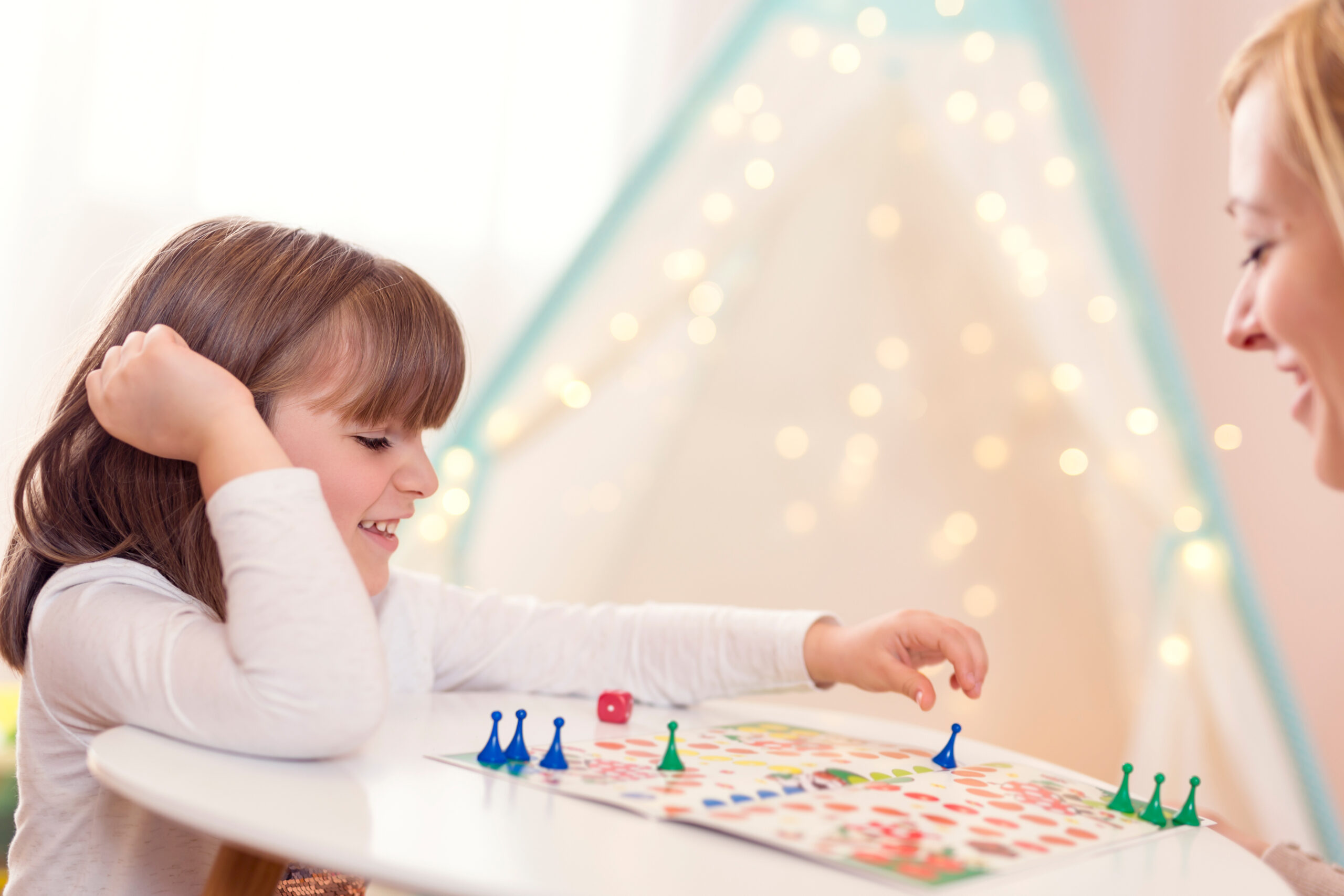 Mother and daughter sitting in a playroom, playing a ludo game; daughter repositioning the pawn